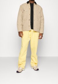 Beige lærredjakke med to forlommer, sort krave, sort t-shirt, gule sweatpants og mønstrede sneakers med lilla accenter.