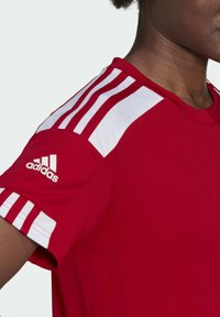 Rotes Sportshirt mit weißen Akzenten. Verfügt über drei weiße Streifen auf der Schulter und ein Adidas-Logo auf dem Ärmel. Glatte Stofftextur.