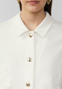 White collared shirt featuring five metallic buttons, smooth texture, and stitched detailing. Gold accents on buttons add distinct detail.