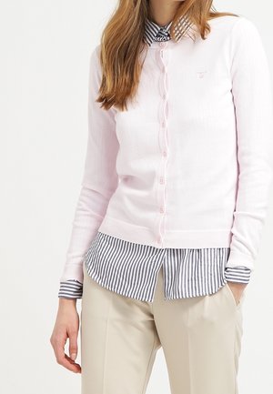 Kobieta ubrana w jasnoróżowy kardigan na guziki, noszony na czarno-białej koszulce w paski, w połączeniu z beżowymi spodniami, trzymająca ręce w kieszeniach.