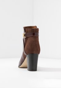 Bottine marron avec une tige en daim, un talon brillant et une bride latérale ornée d'une boucle dorée. Conçue avec un bout arrondi et une hauteur de talon modérée.