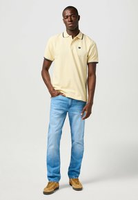 Poloshirt in blassgelb mit marineblauen Akzenten, kombiniert mit hellblauen Jeans und braunen Schuhen. Verfügt über ein kleines Logo auf der linken Brust.