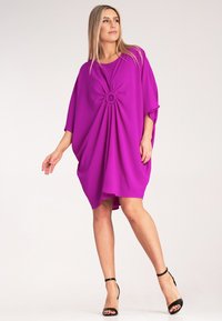 Fioletowa, oversize sukienka z okrągłym węzłem z przodu, krótkimi rękawami i luźnym krojem, w połączeniu z czarnymi wysokimi sandałami na cienkiej szpilce.