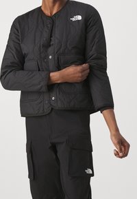 Černá prošívaná bunda s kulatým výstřihem, patentkovým uzávěrem, dvěma předními kapsami a logem The North Face na levém rameni.