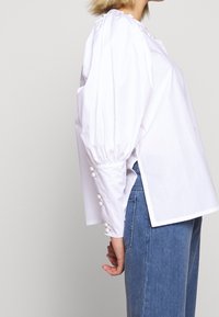 Blusa bianca in cotone con maniche a sbuffo, spacchi laterali e bottoni decorativi in perla lungo i polsini. Abbinata a jeans in denim blu.