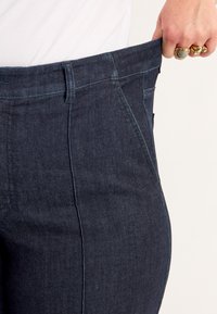 Käsi, millel on kuldsed sõrmused, tõmbab lahtiste tumesiniste teksade vöökohtade külge, mille all on valge topp.