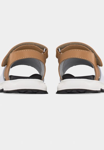 Λευκά και καφέ σανδάλια ανοιχτής πλάτης με μαύρα επενδυμένα πέλματα και χοντρές λευκές σόλες, εμφανιζόμενα από πίσω σε γκρι φόντο.