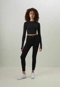 Svart långärmad crop-top med figursydd design, kombinerad med högmidjade svarta skinny jeans och vita sneakers. Modellen står mot en ljus bakgrund.