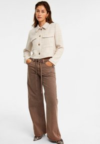 Licht tweed jasje met twee voorkruiszakken, knoopsluiting en opgerolde mouwen, gecombineerd met bruine wijde broekspijpen en lakschoenen met luipaardprint.