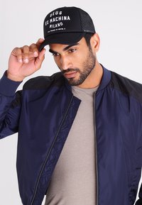 Giacca bomber blu navy con accenti neri sulle spalle, abbinata a un cappellino in rete nero con testo bianco. Lo sfondo è bianco semplice.