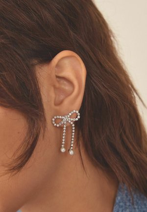 Cercei în formă de fundă din rhinestone, cu lanț suspendat și accente de perle, de culoare argintie, având detalii complexe și o textură netedă.