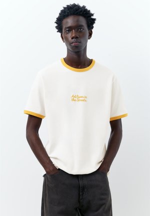 Jeune homme les mains dans les poches, portant un t-shirt blanc avec un liseré jaune et le texte « Art lives in the Streets » sur un fond uni.