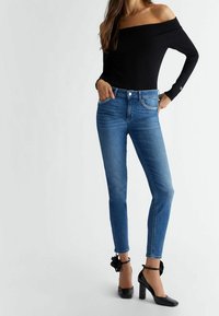 Mujer con blusa negra fuera del hombro, jeans ajustados azules con detalles de strass en los bolsillos y zapatos de tacón negro con tiras en el tobillo.