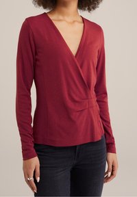 Woman wearing a long-sleeve, deep red wrap-style top and dark jeans, standing against a plain beige background.