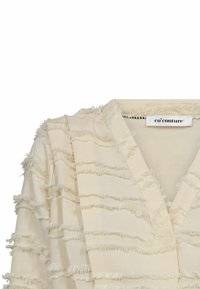 Blusa color crema con un tessuto morbido e testurizzato, con frange cucite. Design a scollo a V con pannelli sovrapposti e un'etichetta visibile all'altezza del collo.