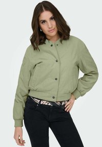 Veste bomber courte vert clair avec boutons-pression, poignets côtelés et deux poches latérales, associée à un jean noir et une ceinture à motif.
