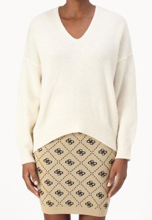 Person iført en cremefarvet V-hals sweater og beige strikket nederdel med sort geometrisk mønster og logoer, stående mod en hvid baggrund.