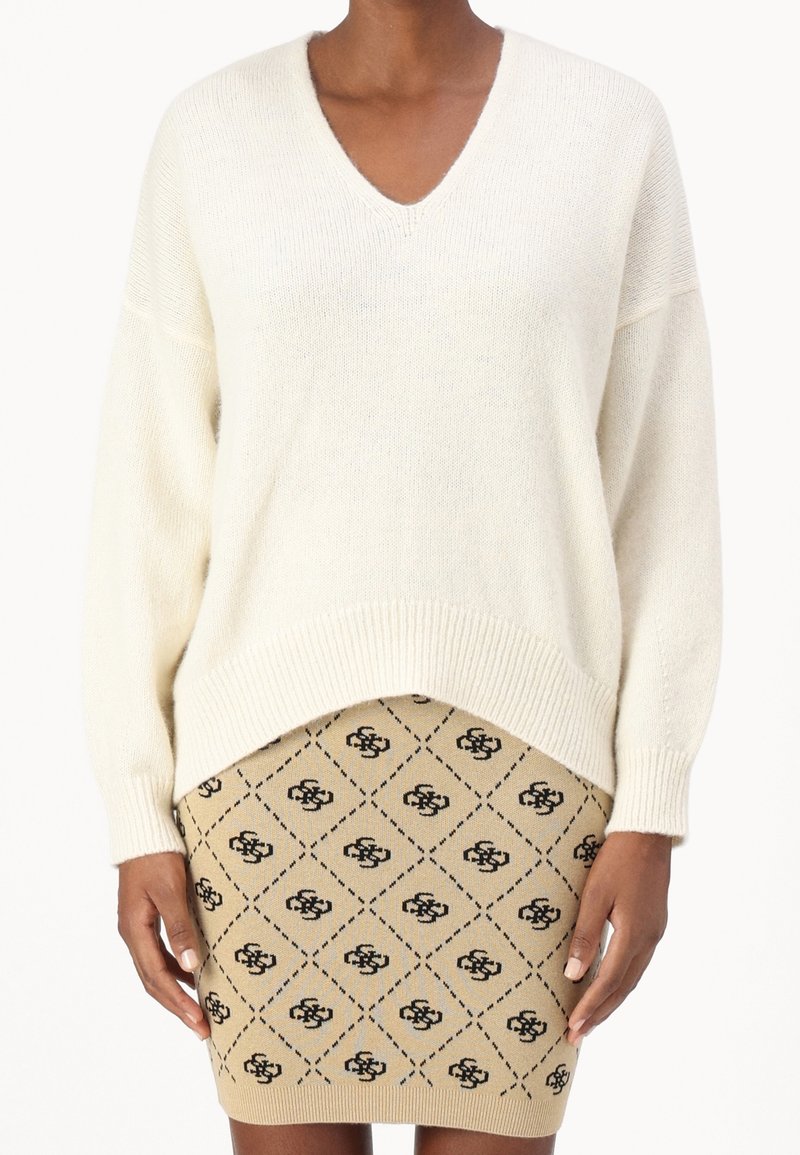 Personne portant un pull à col en V crème et une jupe tricotée beige avec un motif géométrique noir et des logos, debout devant un fond blanc.