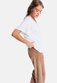 Bílý tričko s krátkým rukávem a véčkovým výstřihem spárované s hnědými manšestrovými kalhotami, které mají vertikální pruhy a volný střih. Viditelná textura přirozené látky.