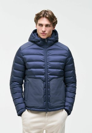 Junger Mann trägt eine marineblaue gesteppte Jacke mit Kapuze, steht mit den Händen in den Taschen und schaut seitlich vor einem einfarbigen Hintergrund.
