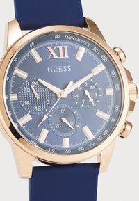 Orologio dorato con quadrante blu, dotato di tre sotto-quadranti, indici delle ore dorati e cinturino in silicone blu. Scala tachimetrica sulla lunetta.