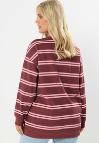 Blondynka odwrócona tyłem, ubrana w długi sweter w paski w kolorze bordowym i różowym oraz jasnoniebieskie dżinsy na jednolitym tle.