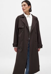 Brun trenchcoat med en stor krage, dubbelknäppt knäppning, långa ärmar och justerbara manschetter, i kombination med denimjeans.