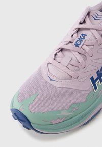 Ανοιχτό μωβ αθλητικό παπούτσι με επάνω μέρος από δίχτυ και συνθετικό υλικό. Πράσινες λεπτομέρειες στη μύτη και τη φτέρνα, υφή επιφάνειας, μπλε λογότυπο και επίπεδα κορδόνια.