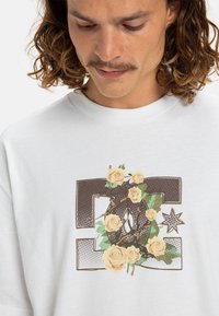 Camiseta blanca de manga corta con un diseño gráfico que presenta rosas beige y una letra "D" de color marrón con un patrón. La tela parece suave y casual.