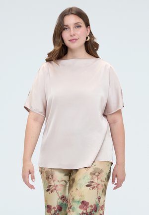 Kvinde med bølget brunt hår iført en lysebeige kortærmet bluse og beige bukser med blomsterprint, stående foran en ensfarvet baggrund.