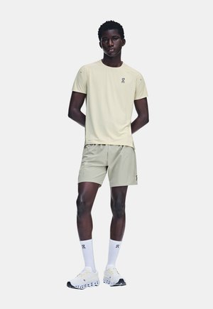 Jeune homme debout les mains dans le dos, portant un t-shirt de sport beige clair, un short assorti, des chaussettes blanches et des chaussures de course blanches.
