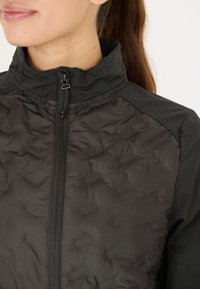 Schwarze Zip-Jacke mit gep quilteter Textur auf der Vorderseite und glattem Stoff an den Schultern. Hoher Kragen mit Reißverschluss.
