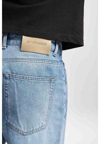 Ljusblå jeans i denim med en läderetikett på midjebandet där det står "2Y STUDIOS." Jeansens design inkluderar en bakficka och en klassisk skärning.