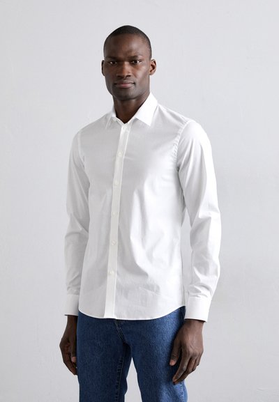 Camicia bianca a maniche lunghe in cotone, con colletto appuntito, chiusura con bottoni e vestibilità sagomata. Abbinata a jeans di denim blu.