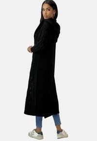 Svart långstickad cardigan med huva, med kabelstickad struktur och ribbade kanter, matchad med ljusblå jeans och vita sneakers.