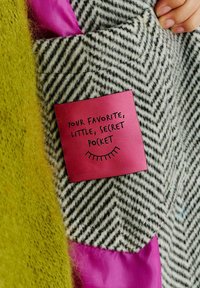 Houndstooth látka v černobílé barvě s růžovou etiketou s nápisem "Vaše oblíbená, malá, tajná kapsa" a zeleným texturovaným akcentem.