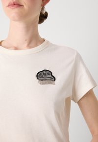 Cream cotton t-shirt featuring a black cloud patch with silver fringe embellishment on the left chest. Short sleeves and round neckline.