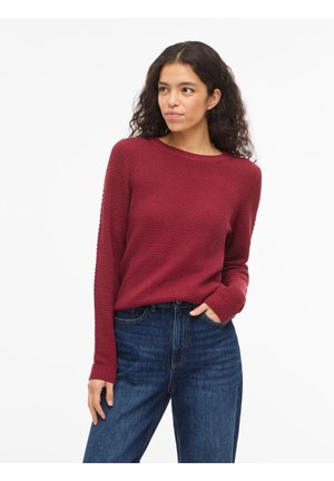 Femme aux longs cheveux bouclés portant un pull bordeaux texturé et un jean taille haute bleu, une main dans la poche, regardant sur le côté.