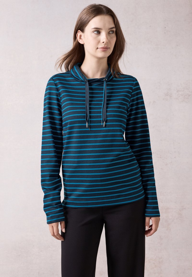Maglietta a maniche lunghe con cappuccio di colore blu scuro con strisce orizzontali color turchese, dotata di un collo con cordoncino e vestibilità comoda. Abbinata a pantaloni neri.