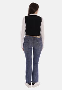 Gilet nero corto sopra una camicia bianca, abbinato a jeans a zampa d'elefante blu. I jeans presentano tasche posteriori e un'etichetta sulla vita. Sneakers bianche completano l'outfit.