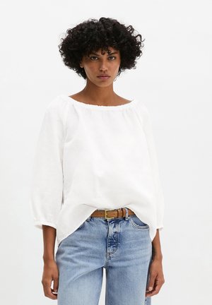 Donna con capelli ricci scuri che indossa una camicetta bianca larga a maniche lunghe, jeans blu a vita alta e cintura marrone su uno sfondo bianco uniforme.