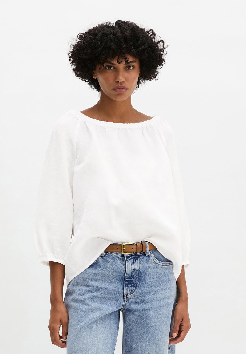 Femme aux cheveux bouclés foncés portant un chemisier blanc ample à manches longues, un jean bleu taille haute et une ceinture marron sur un fond blanc uni.
