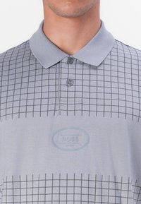 Graues Polo-Shirt mit Karomuster, klassischem Kragen, zwei Knöpfen am Hals und einem strukturierten Logo-Patch in der Mitte.