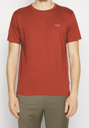 Mężczyzna w jednolitej, rdzawoczerwonej koszulce z krótkim rękawem z logo „JOOP! JEANS” na piersi oraz oliwkowo-zielonych spodniach, stojący na białym tle.