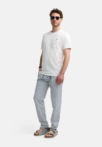 Hvid T-shirt med korte ærmer og lomme; blå- og hvidternede bukser; grå sandaler med to stropper; model iført sorte solbriller.