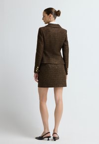 Žena s drdolem, oblečená v hnědém texturovaném blejzru a odpovídající sukni, zezadu, stojící na bílém pozadí s černými sandály na podpatku.