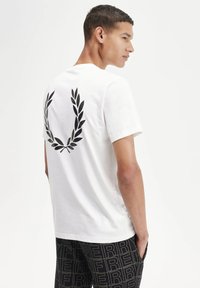 Camiseta de algodón blanca con un diseño de corona de laurel negra en la parte posterior. La camiseta tiene un cuello redondo y mangas cortas.