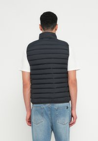 Pudret sort vest med lodret quiltet design, høj krave og letvægtsmateriale, båret over en hvid t-shirt og blå jeans.