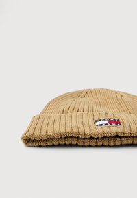 Beanie in maglia a coste in beige con risvolto, caratterizzato da un piccolo patch rettangolare in rosso, bianco e blu su uno sfondo uniforme.