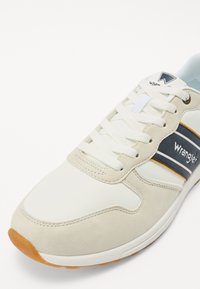 Fehér sneaker bézs suede díszítéssel, sötétkék csíkkal és "Wrangler" márkajelzéssel. Fűzőkkel és texturált gumitalppal rendelkezik.